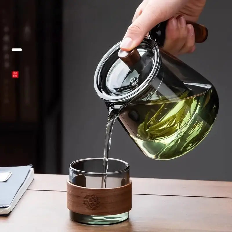 A clear glass tea set with wooden handle teapot pouring green tea on a retro glass tray with bamboo cup sleeve