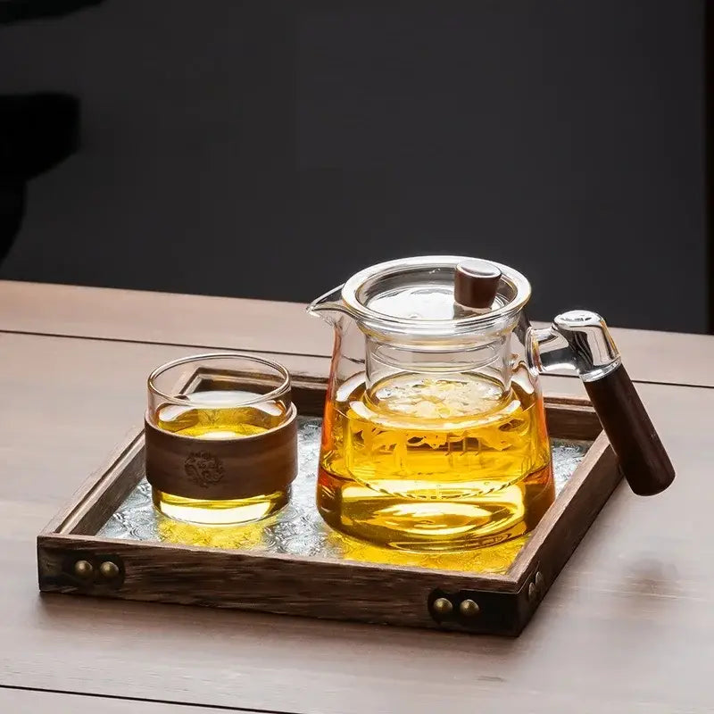 Clear glass teapot with wooden handle and lid knob filled with golden tea on a retro glass tray tea set by DecorBites