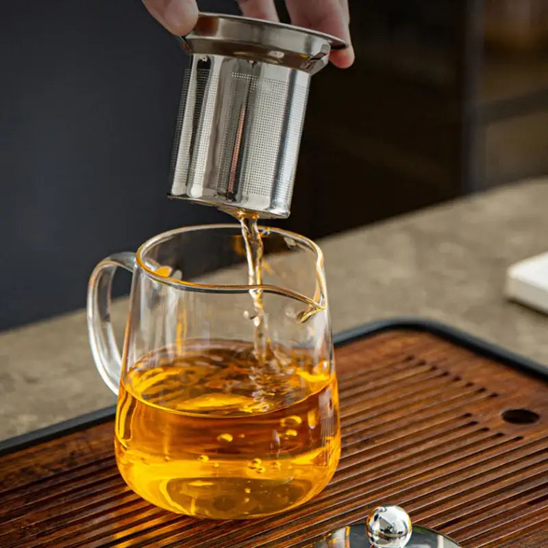A stainless steel tea infuser is lifted from a clear decorbites glass teapot with golden brewed puer tea flowing below