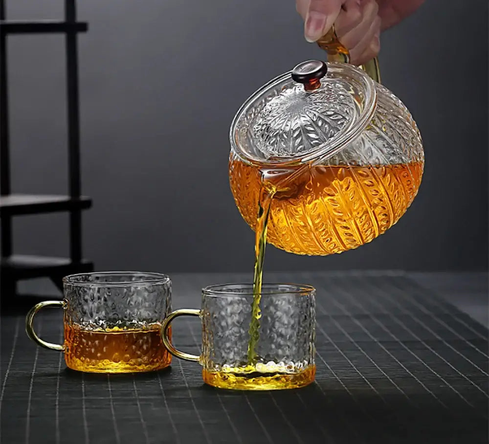 Clear glass decorbites glass teapot with embossed patterns and black knob pouring amber tea into textured mugs