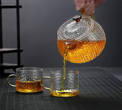 Clear glass decorbites glass teapot with embossed patterns and black knob pouring amber tea into textured mugs