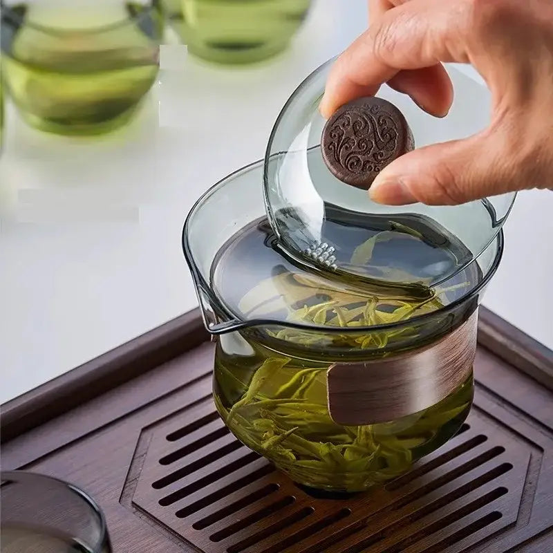 A hand lifts a dark brown engraved lid from a decorbites glass teapot set filled with green tea leaves