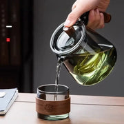 Clear glass decorbites glass teapot set with wooden handle pouring green tea into glass cup with bamboo sleeve