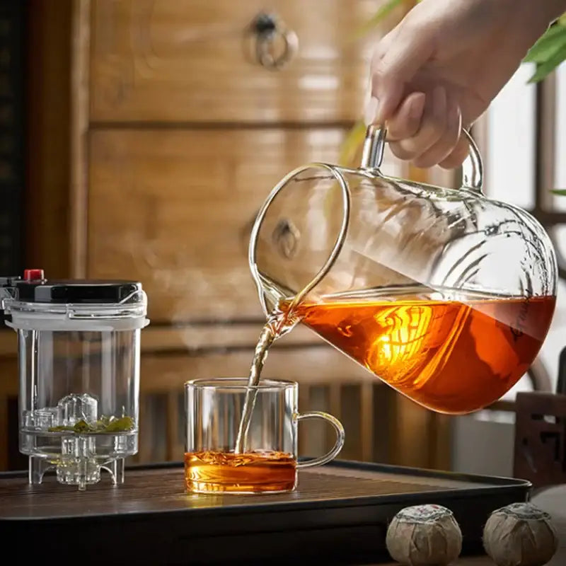 A clear glass decorbites glass teapot with one-button filter pouring amber tea into a matching glass cup