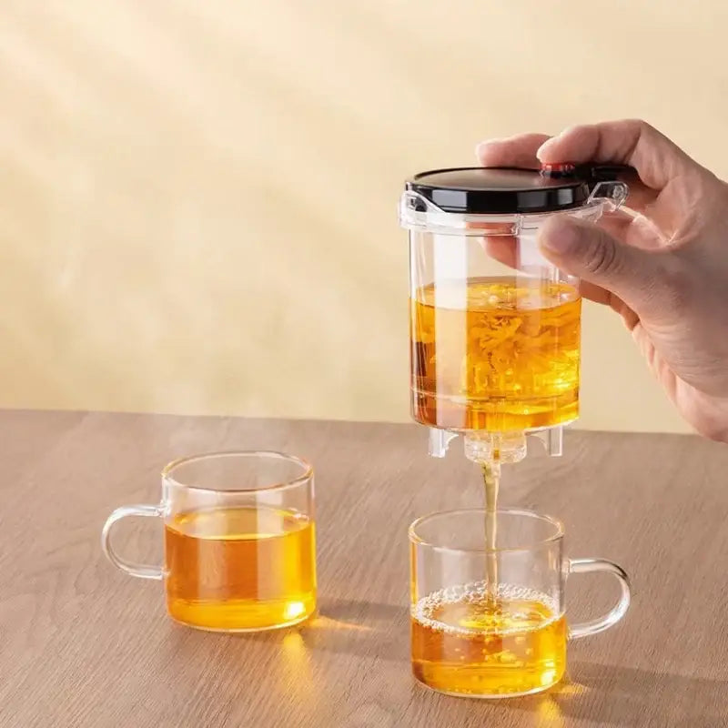 Clear glass decorbites glass teapot with black lid pouring golden tea into transparent mugs for tea brewing
