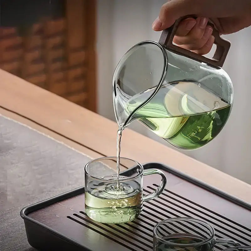 Clear glass decorbites glass teapot with dark brown wood handle pouring green tea into matching glass cup for kung fu tea ceremony