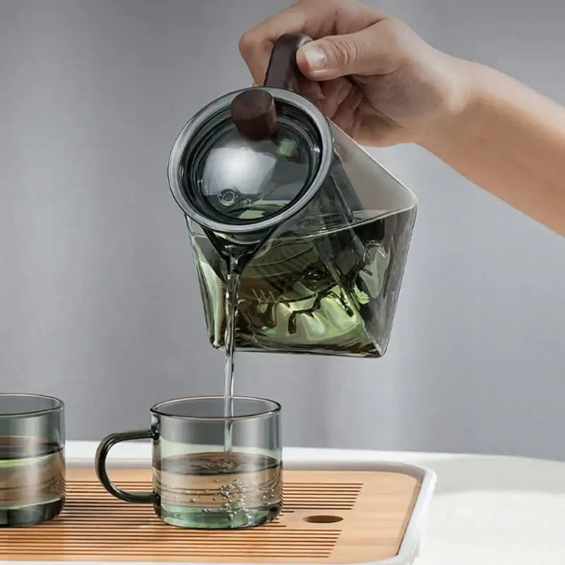 A sleek dark green decorbites glass teapot with wooden handle pouring tea into a matching clear glass mug