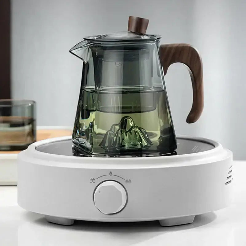 A decorbites glass teapot with a transparent body and dark wood handle sits on a white electric heating base