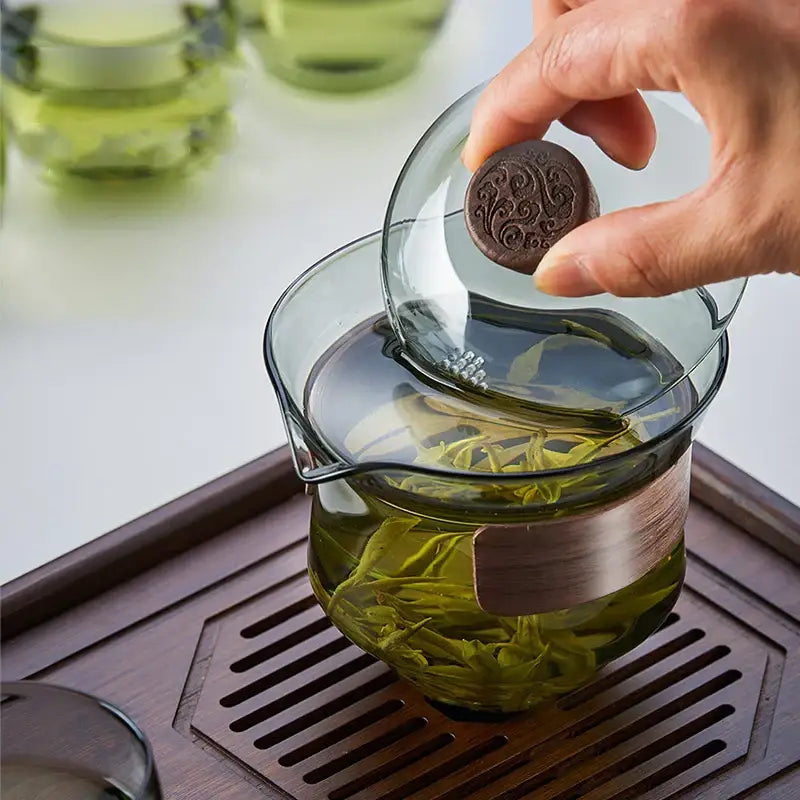 A clear glass DecorBites glass tureen teapot with a dark brown engraved lid lifted by hand for kung fu tea set use