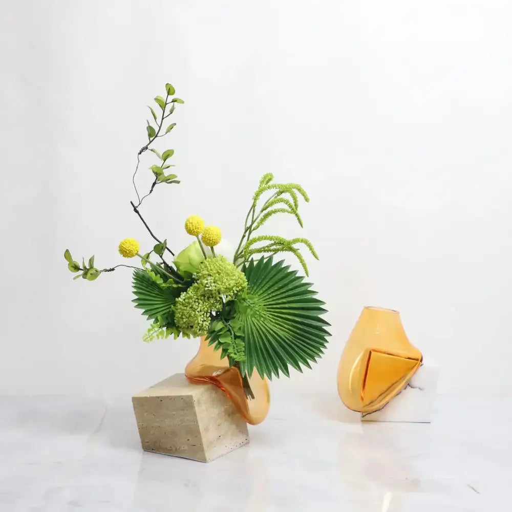 A modern amber DecorBites glass vase with a marble base holds green foliage and yellow flowers for elegant décor
