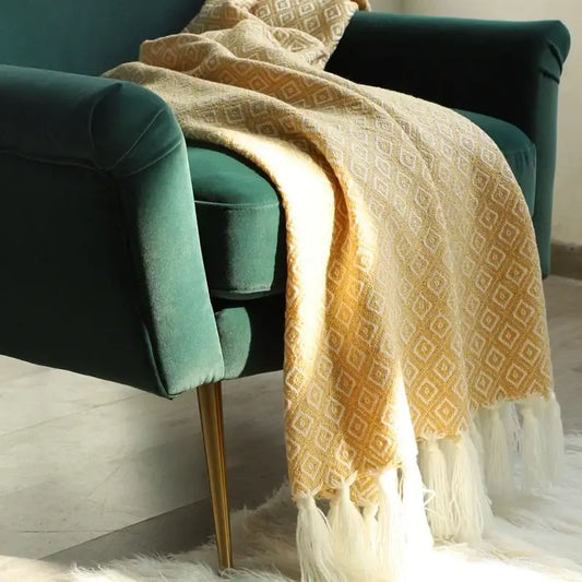 A cream decorbites golden knitted blanket with geometric pattern and white fringe draped over a green velvet chair
