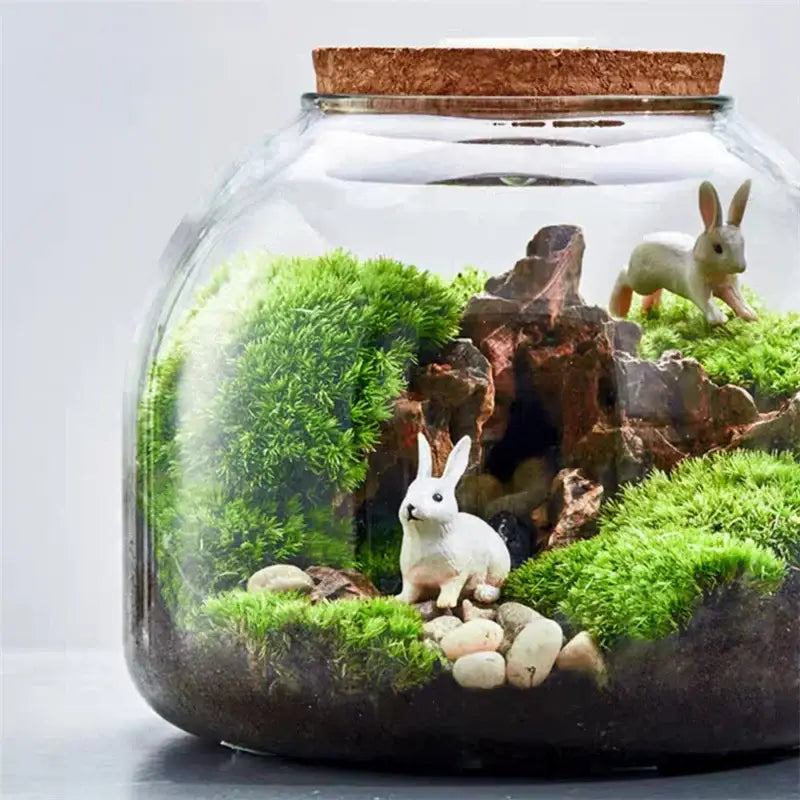 A clear glass vase terrarium with green moss and white rabbit figurines in the decorbites green moss glass container
