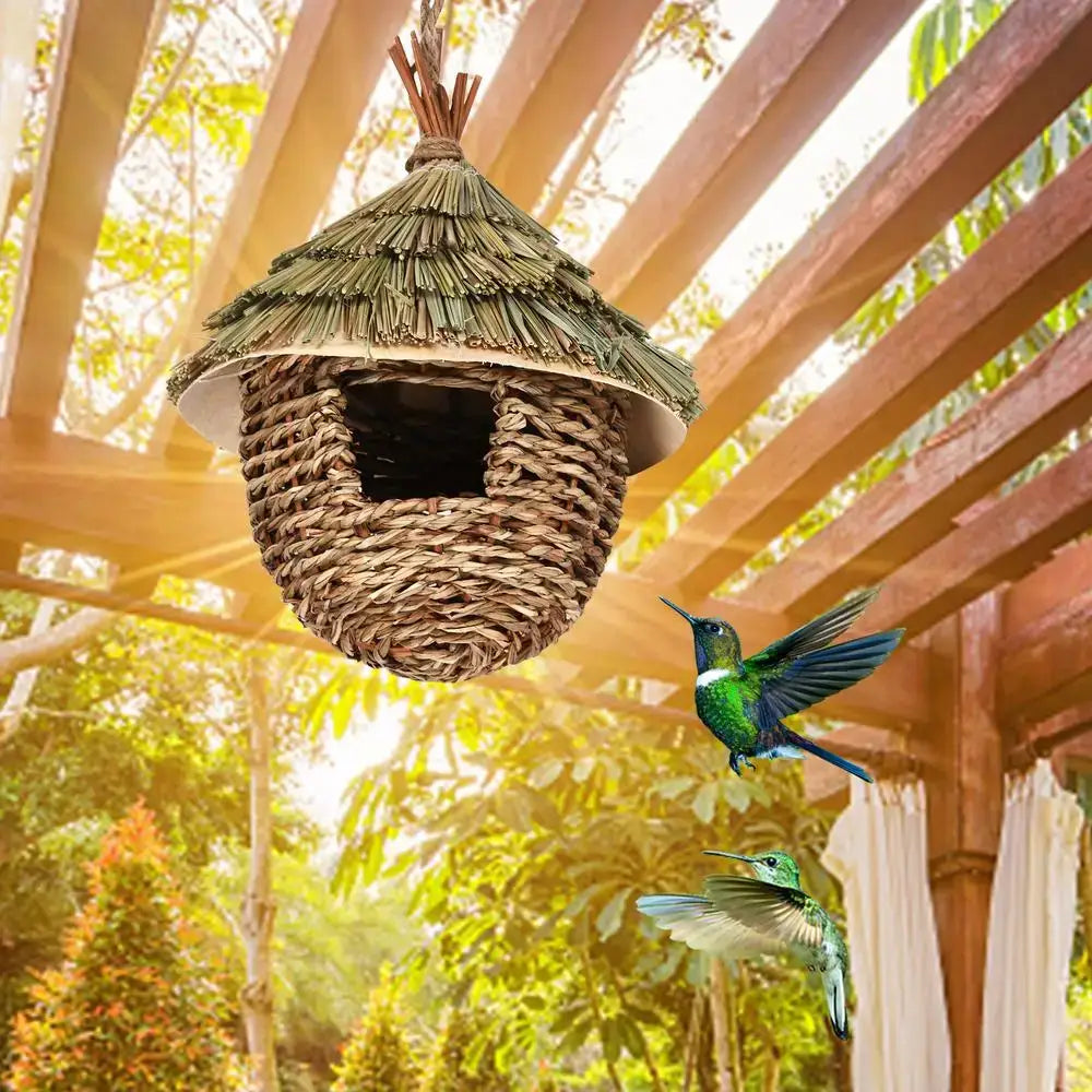 A rustic hand-woven hummingbird house with thatched roof and natural brown tones hangs from a wooden pergola