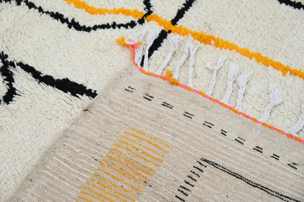 Cream-colored moroccan azilal rug with bold black and orange geometric patterns and vibrant pink-accented white fringe
