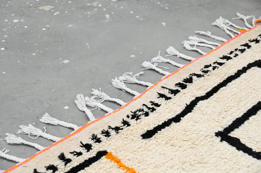 Cream-colored moroccan azilal rug with bold black geometric patterns vibrant orange border and long white fringe 240x152 cm