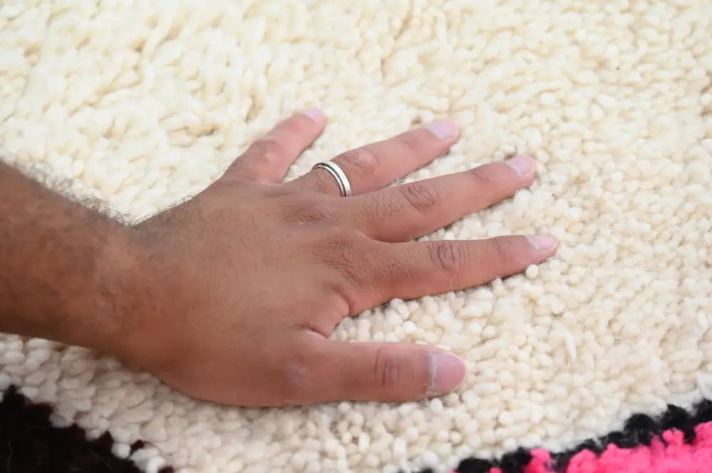 A hand with a silver ring resting on a cream azilal rug with bold black and pink border made of natural wool 250x160cm