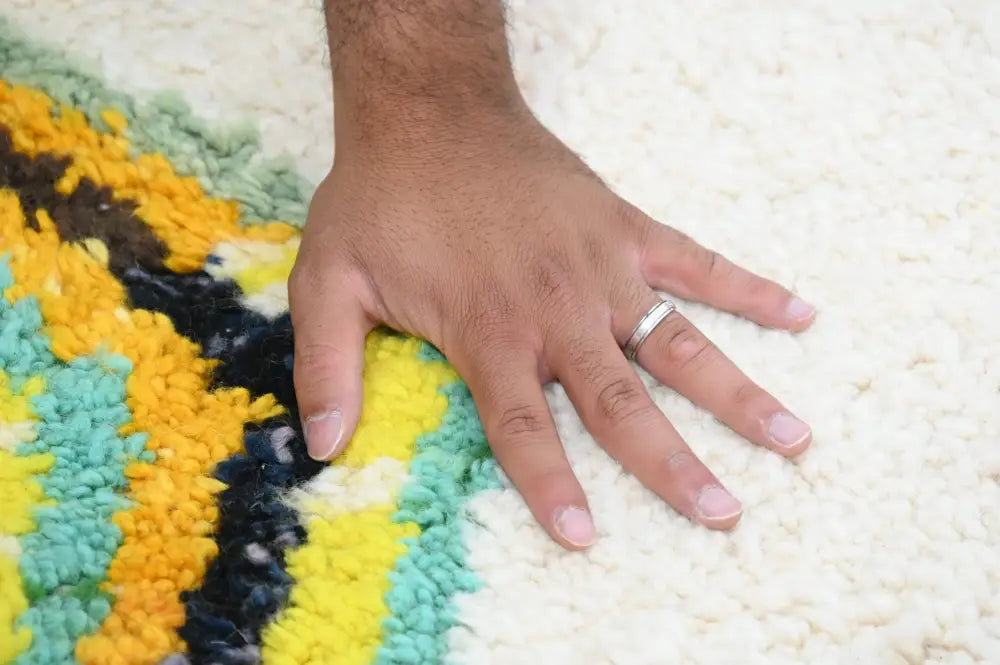 A hand rests on a handmade moroccan azilal rug with bold yellow teal and black stripes on creamy white background