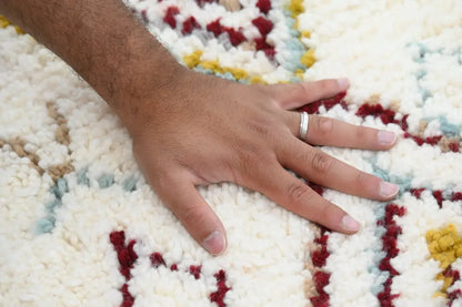 A hand with a silver ring rests on a plush cream handmade moroccan azilal rug with red and yellow geometric patterns