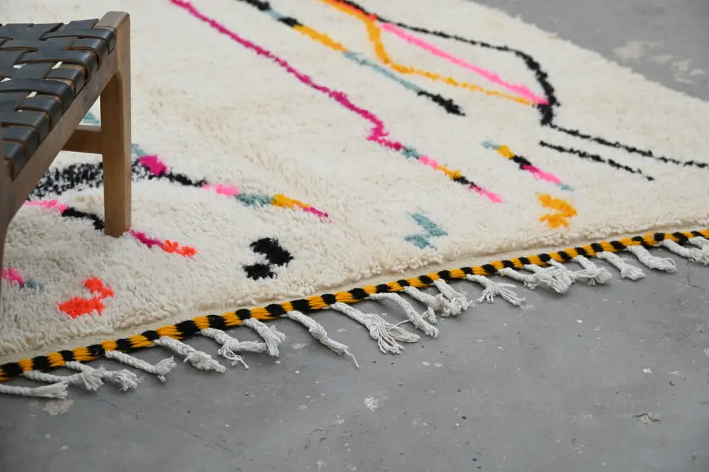 A fluffy off-white handmade moroccan azilal rug with vibrant pink yellow black geometric patterns and striped tassel border