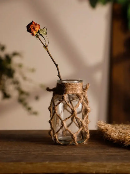 A clear hanging glass vase with hemp rope and woven jute wrap holding a single dried orange rose for rustic decor