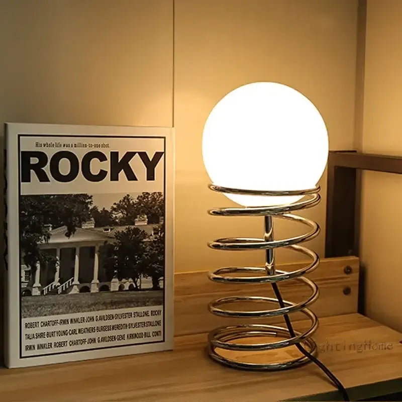 A sleek ins bauhaus spring table lamp with a glowing white glass globe on a polished chrome spiral spring base