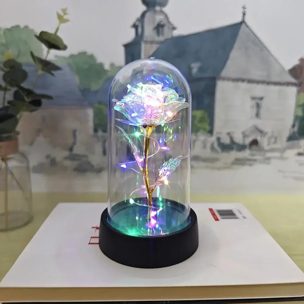A glowing iridescent eternal rose light with a golden stem inside a clear glass dome on a black base by decorbites