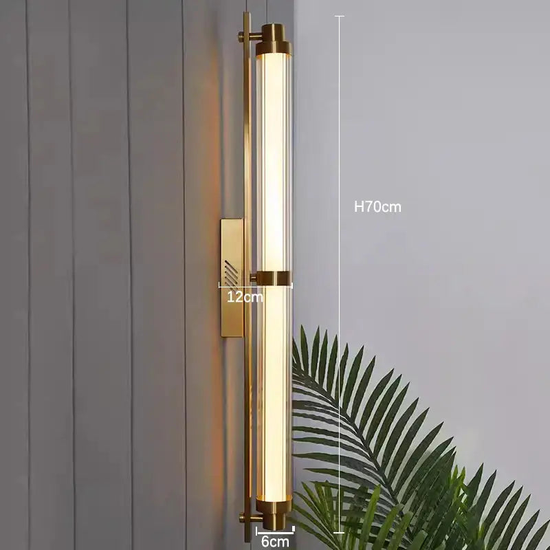 A tall slender brass decorbites led glass wall lamp with a clear cylindrical tube emitting warm light for living spaces