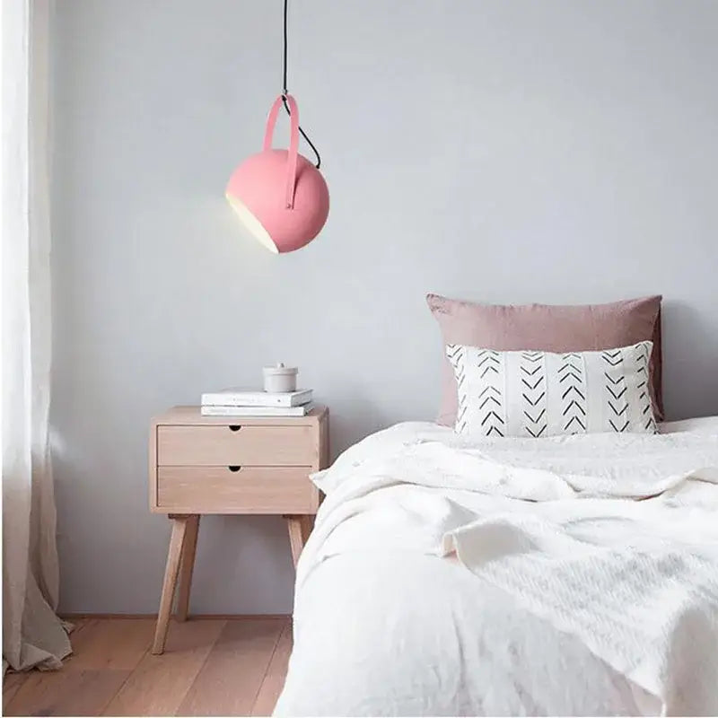 A soft pink spherical DecorBites LED pendant light with black cord hanging above a bed for home decor lighting