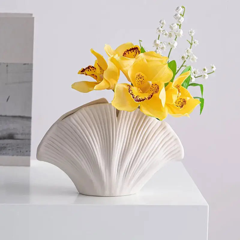 A white decorbites lotus leaf ceramic vase shaped like a scallop shell holding yellow orchids and lily-of-the-valley flowers
