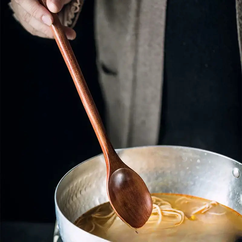 A smooth rich brown decorbites lotus wooden spoon with a long handle held above a pot of soup for cooking use