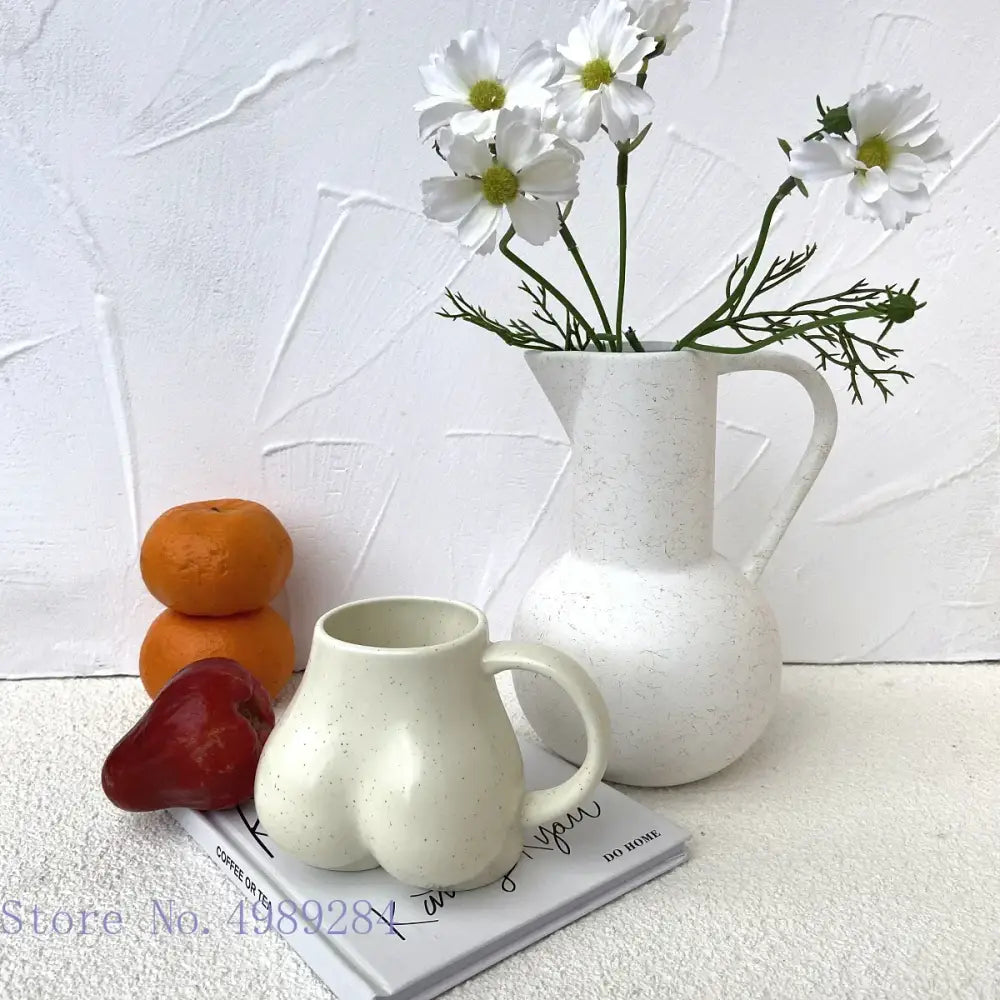White speckled ceramic coffee cup shaped pitcher with handle and spout holding white daisies by DecorBites™