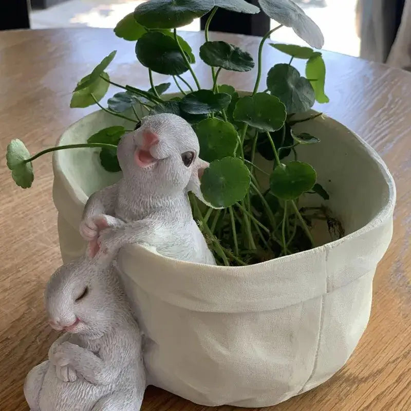 White resin lovely rabbit garden figurines beside a green plant in an off-white ceramic rabbit garden flowerpot
