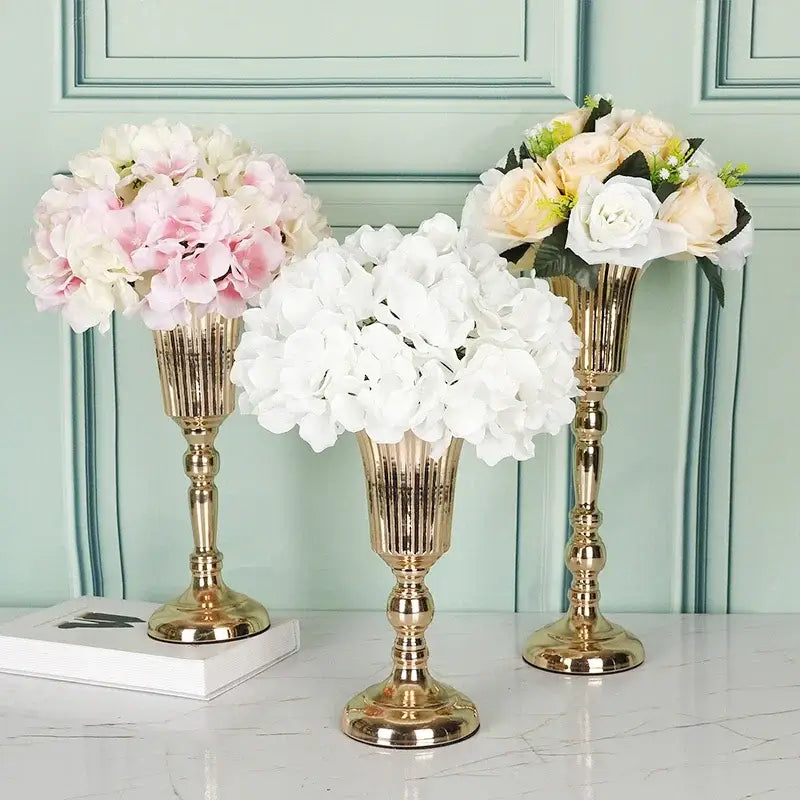 Three elegant gold-toned metal trumpet vases with fluted designs holding soft pink white and cream artificial flowers