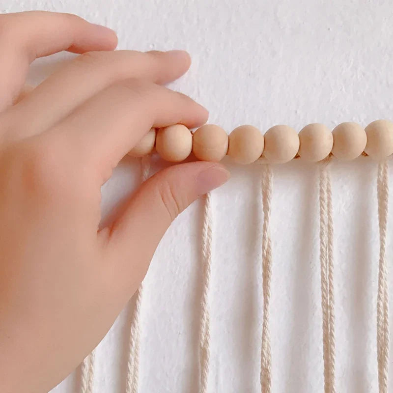 A hand gently holds light beige wooden beads on cream macrame cords in moroccan bohemian wall decor tapestry