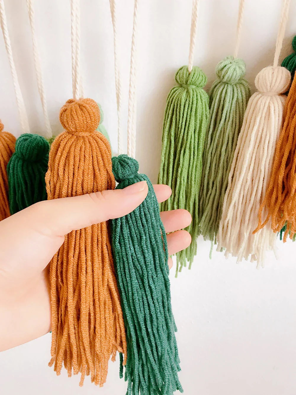 A hand holding two colorful yarn tassels in orange and green for moroccan bohemian wall decor tapestry