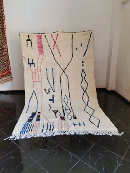 Cream-colored moroccan handmade azilal berber carpet with black pink and blue geometric patterns and fringed edges supporting sustainable practices