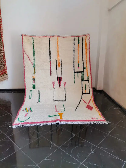 A cream-colored moroccan handmade azilal berber carpet with vibrant green orange black and pink geometric patterns and fringed edge