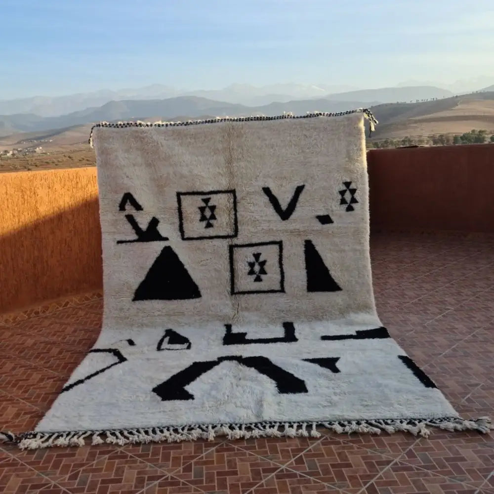 A cream-colored beni ouarain rug with bold black geometric patterns and fringed edges made of moroccan wool