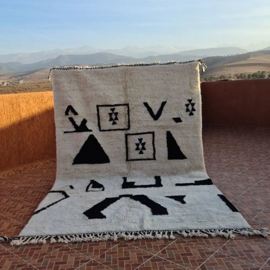 A cream-colored beni ouarain rug with bold black geometric patterns and fringed edges made of moroccan wool
