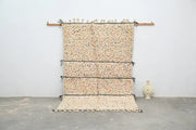Beige moroccan handmade traditional azilal rug with colorful speckles and black bands hanging on wooden rod by decorbites