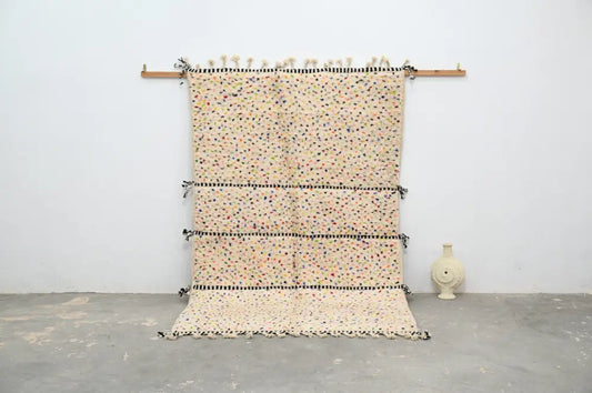 Beige moroccan handmade traditional azilal rug with colorful speckles and black bands hanging on wooden rod by decorbites