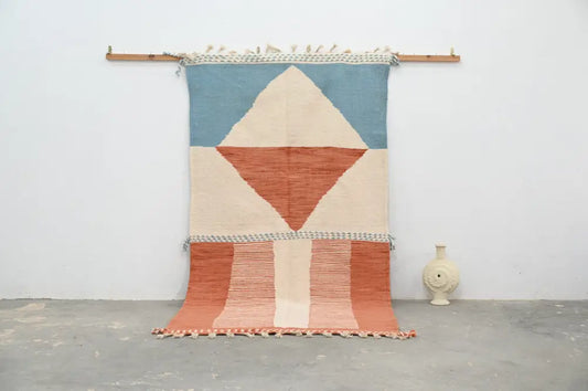 Moroccan handmade traditional azilal rug in soft blue terracotta and cream tones with fringed edges hanging on wooden rod