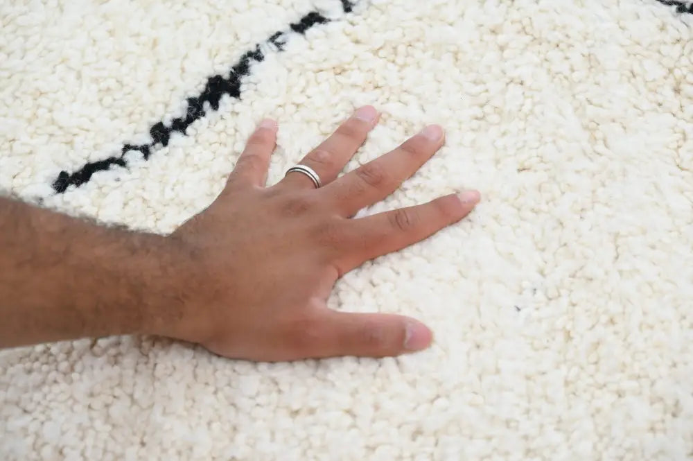 A hand with a silver ring rests on a plush off-white moroccan handmade beni ouarain carpet with black geometric stripes