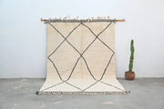 Cream-colored beni ouarain rug with black geometric diamond patterns and fringed edges hanging on wooden rod