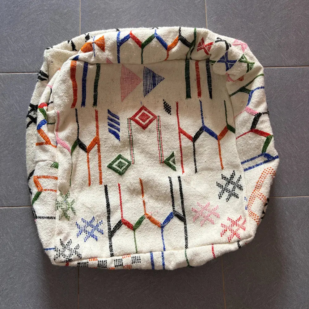 A cream-colored decorbites moroccan handmade pouf 60cm x 60cm with vibrant geometric pattern in red blue green orange and black