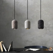 Three cylindrical cement pendant lamps in gray white and black with speckled finish hang against a textured wall industrial style