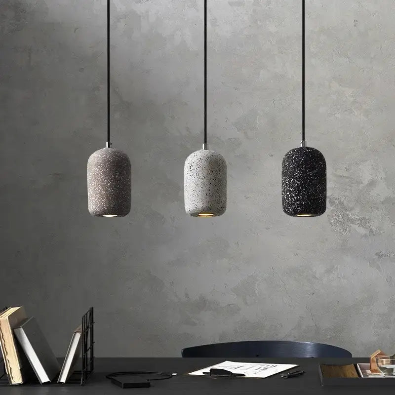 Three cylindrical cement pendant lamps in gray white and black with speckled finish hang against a textured wall industrial style
