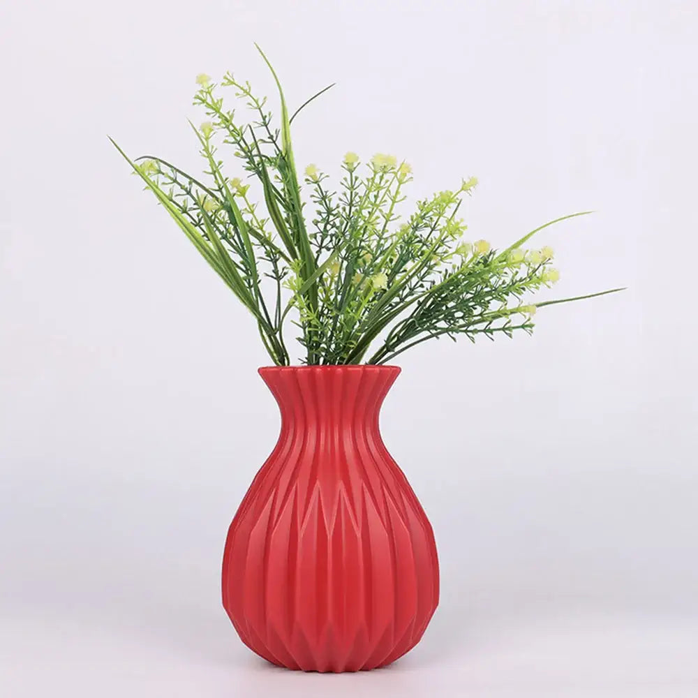 A vibrant red plastic flower vase with geometric ribbed design holds artificial greenery and pale yellow flowers