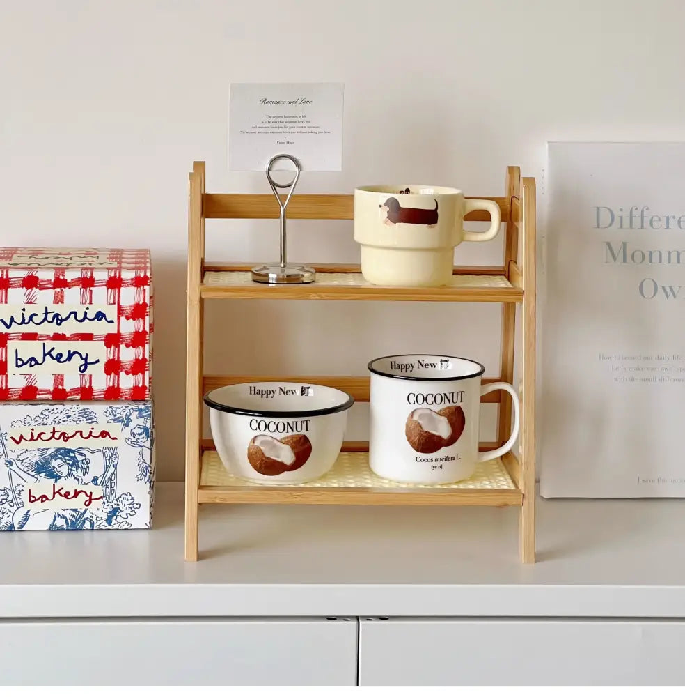 Cream-colored enamel mug with black rim and coconut illustration placed on decorbites rattan 2-tier desktop organizer shelf