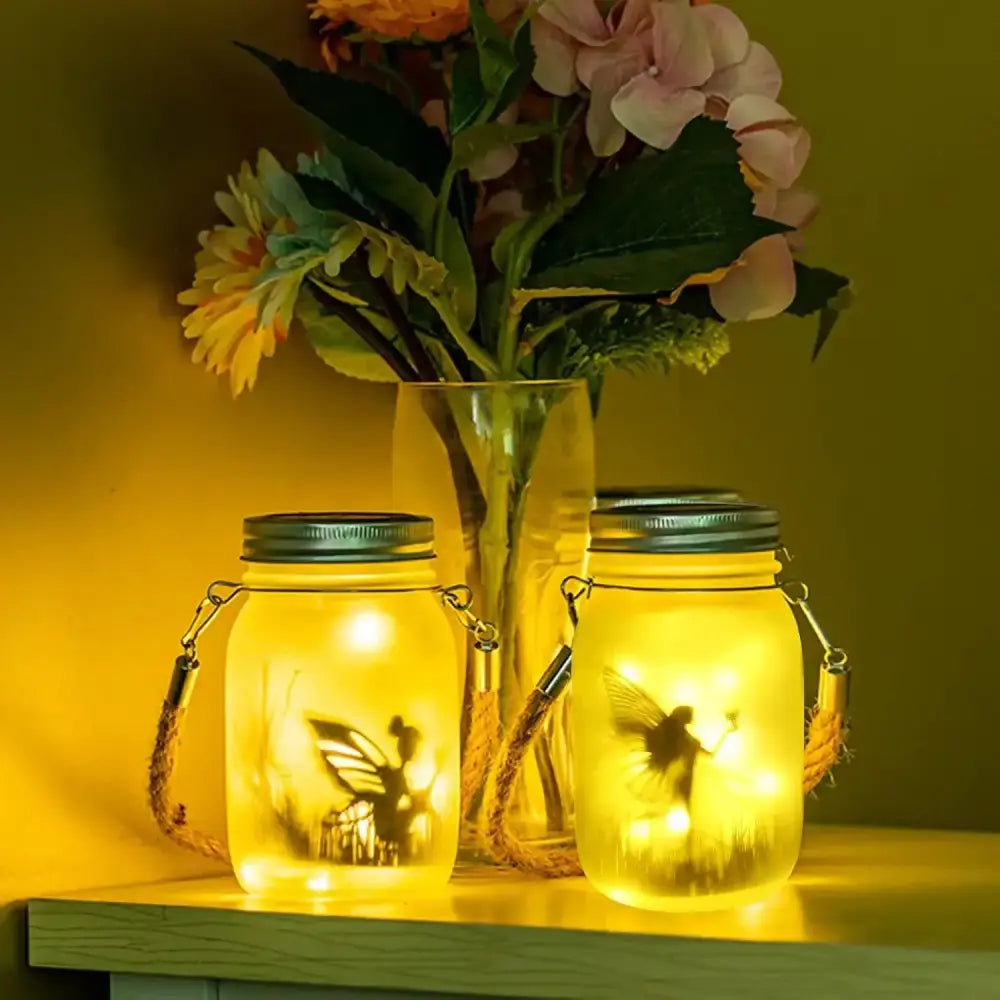 Two illuminated glass solar mason jar lanterns with fairy silhouette cutouts and rope handles for garden decoration